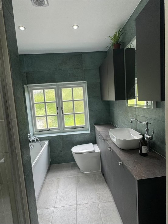 Bathroom with green-grey stone-effect tiles, a white bath, toilet and a grey vanity with a counter-top basin