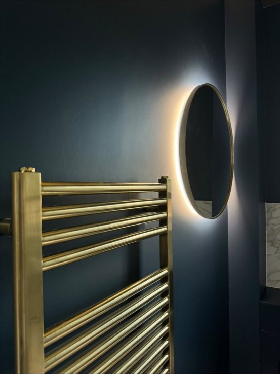 Brushed brass heated towel rail and a backlit round mirror on a deep navy bathroom wall